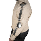 A side profile of a person wearing a beige quilted historical-style jacket with a dark leather shoulder strap and polished metal chain detailing, paired with black pants, against a plain white background.