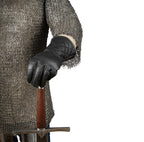 A close-up of a person wearing a chainmail shirt and a black quilted leather glove, holding the pommel of a sword against a plain white background.