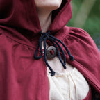 Close-up of a person wearing a burgundy hooded cloak with a black rope tie and a round metal clasp, layered over a cream blouse. The background is a soft, blurred green outdoor setting.