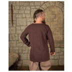 Man wearing brown canvas tunic standing in front of brick wall.