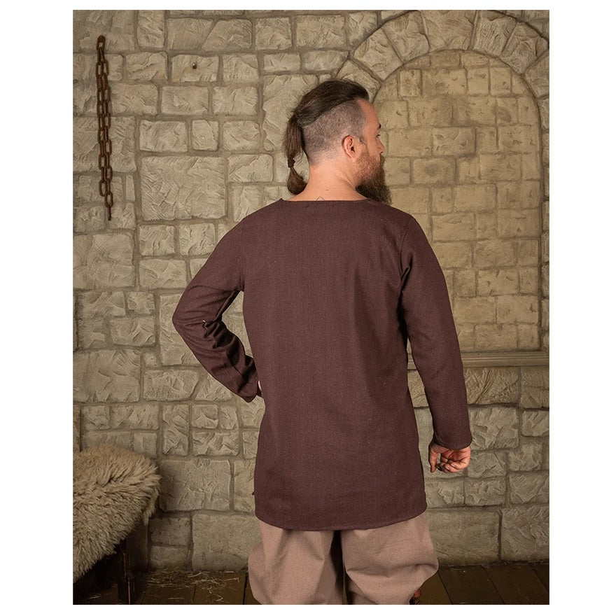 Man wearing brown canvas tunic standing in front of brick wall.