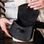 A close-up of a person holding open a black leather pouch or satchel attached to a beige linen tunic with a black belt. The pouch has visible stitching, a leather loop, and a dark interior.