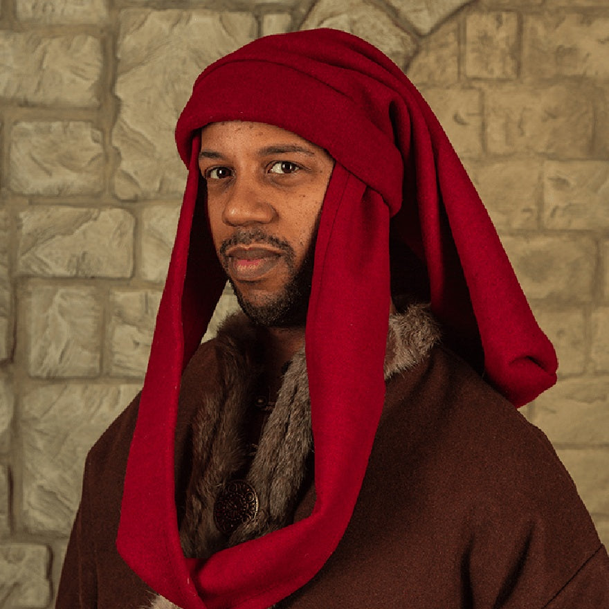 A man wearing a medieval-style costume with a deep red draped head covering and a brown robe with faux fur trim, set against a stone wall background.