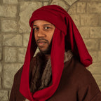 A man wearing a medieval-style costume with a deep red draped head covering and a brown robe with faux fur trim, set against a stone wall background.
